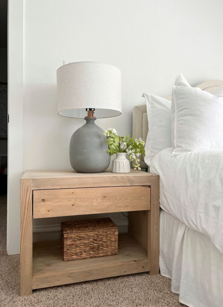 a nightstand with a lamp on top of it next to a white bed and pillows