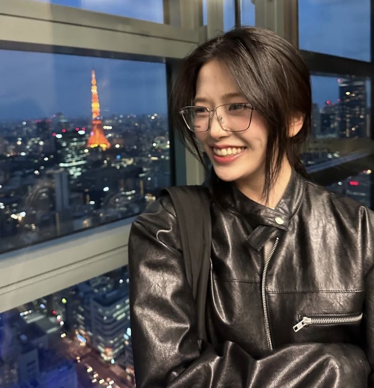 a woman wearing glasses sitting in front of a window with the city lights behind her