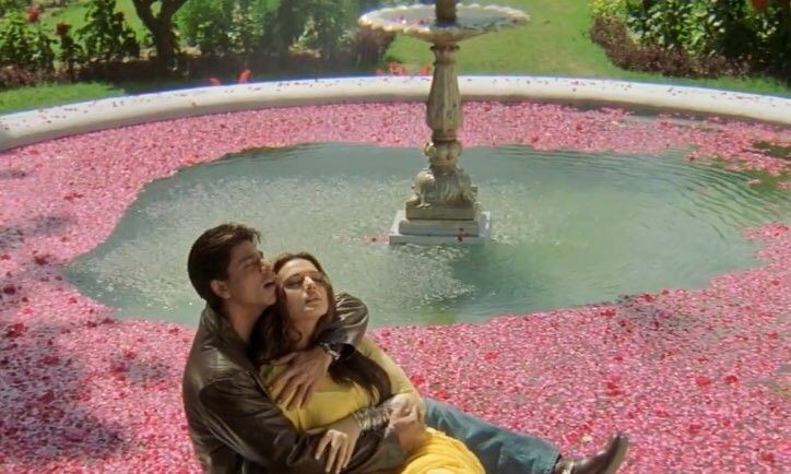 a man and woman sitting in front of a fountain with pink flowers on the ground