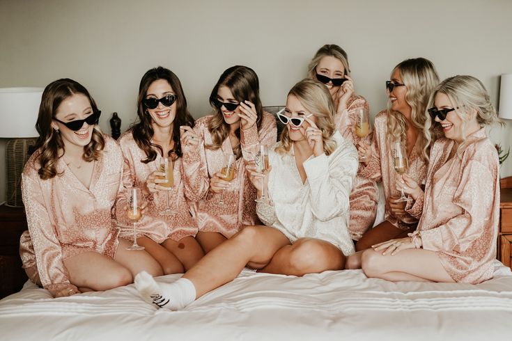 a group of women sitting on top of a bed wearing pink robes and holding champagne glasses