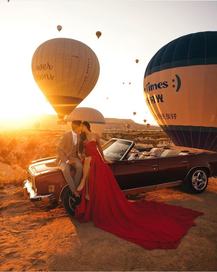 a man and woman sitting on top of a car next to hot air balloons