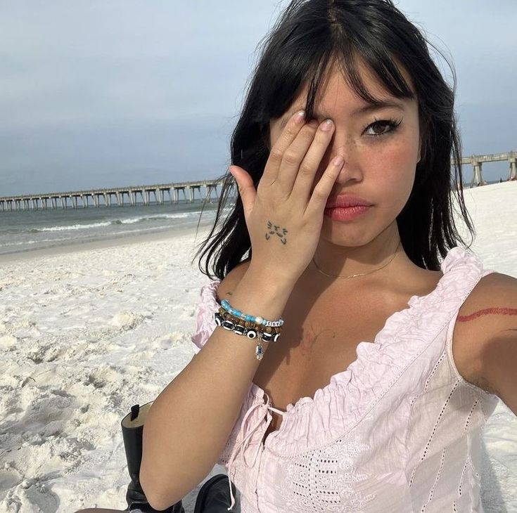 a woman in pink shirt holding her hand up to her face on beach with pier in background