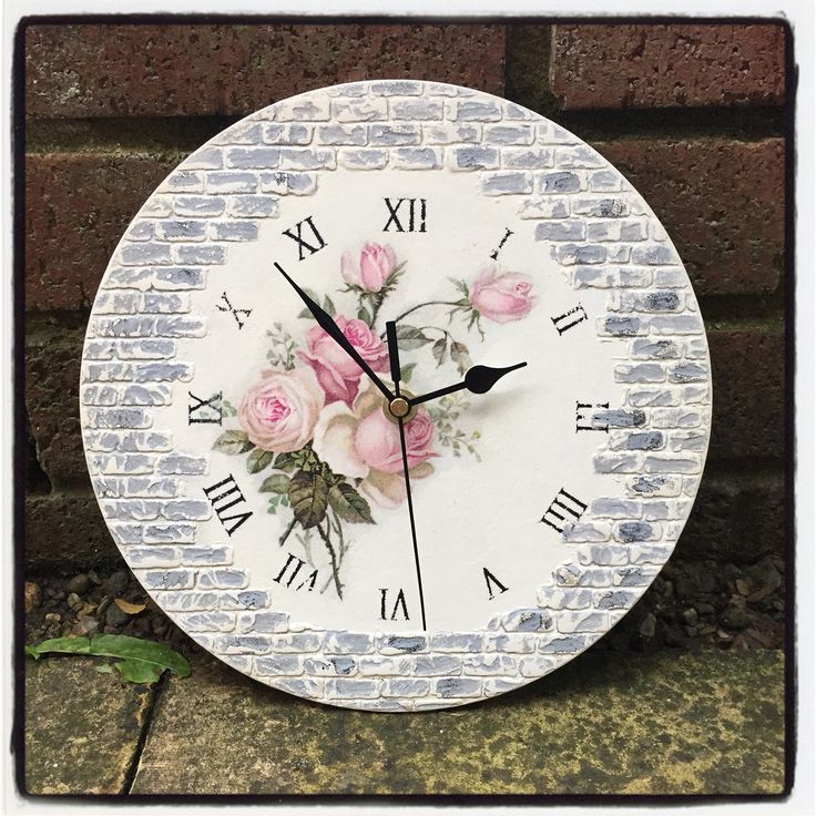 a white clock with pink roses on it sitting next to a brick wall and green leaves