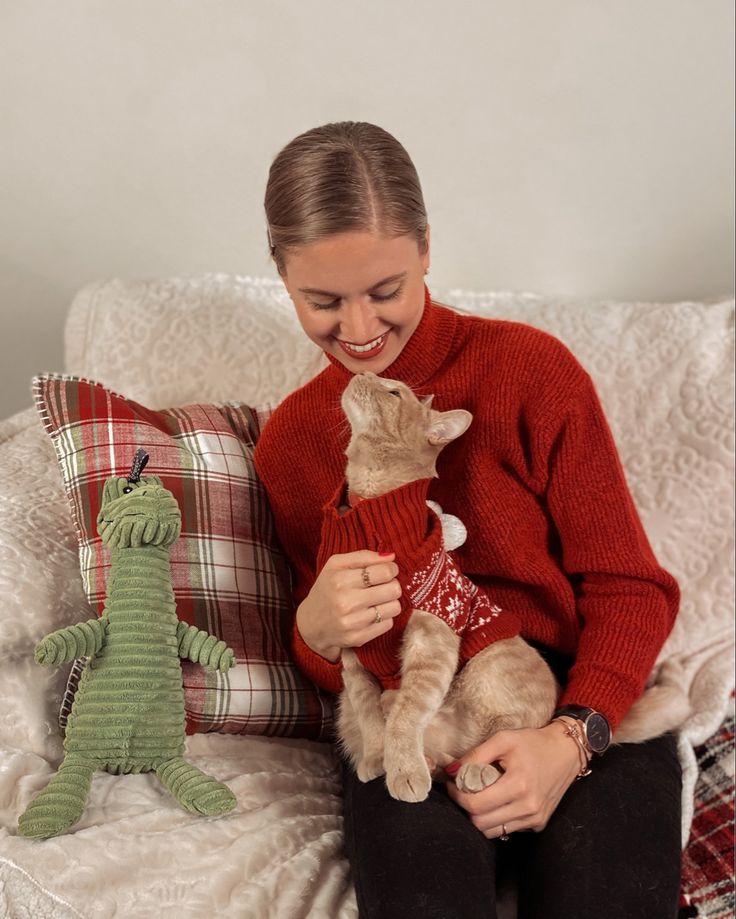 a woman sitting on a couch holding two cats and a stuffed animal toy in front of her