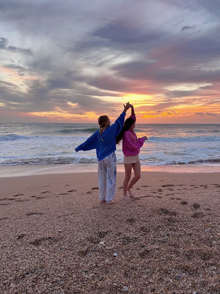 two people are standing on the beach with their arms in the air as the sun sets