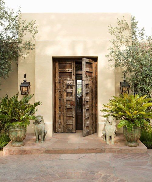 the entrance to an adobe style home with potted plants on either side and large wooden doors