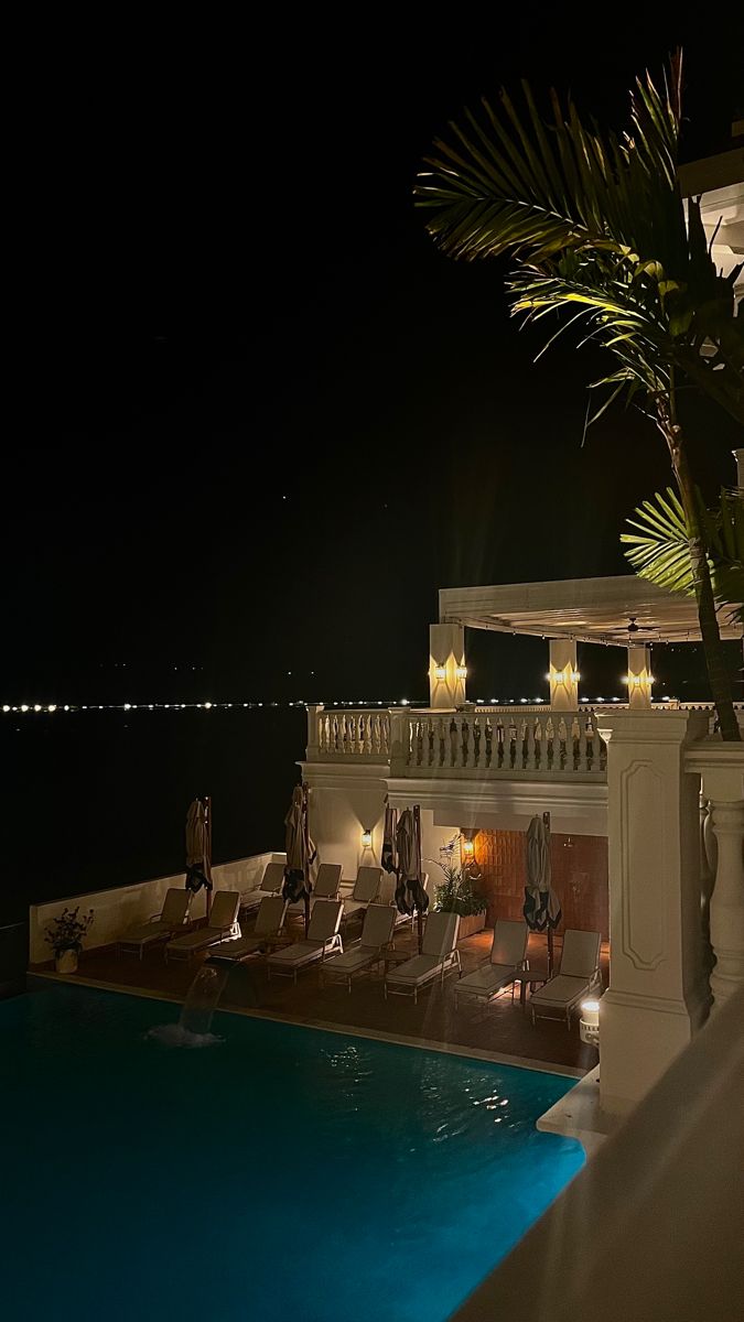 an outdoor swimming pool at night with lounge chairs and palm trees in the foreground