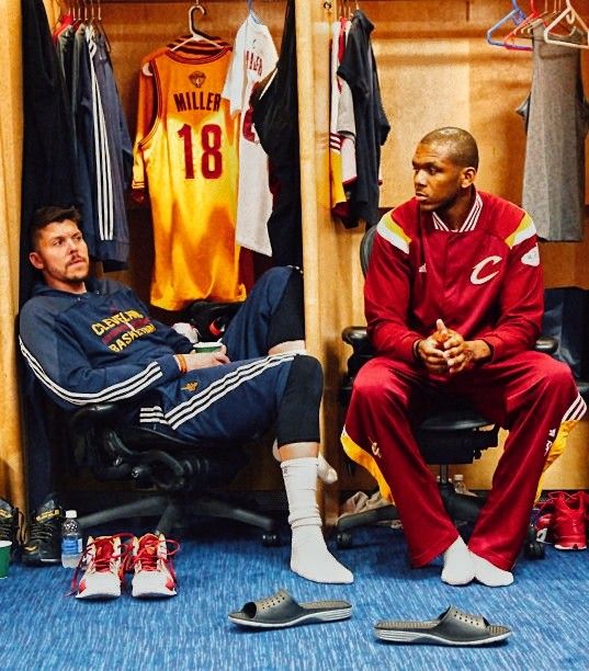 two men sitting in a locker room with shoes on the floor and shirts hanging up