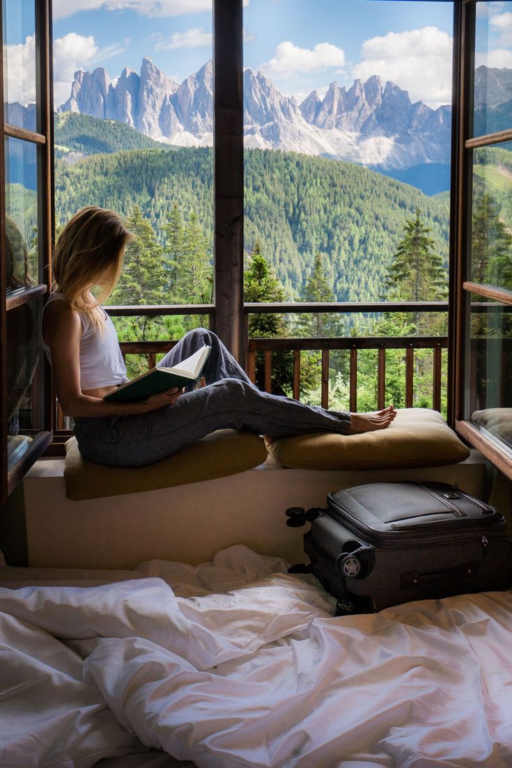 a woman sitting on top of a bed next to a window with mountains in the background