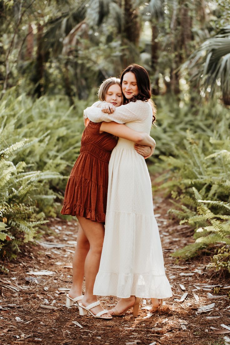 two women hugging each other while standing in front of some trees and plants with their arms around one another