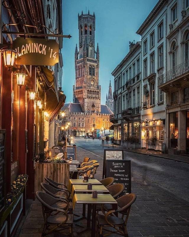 an empty street with tables and chairs in front of a tall clock tower at dusk