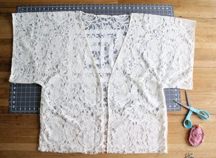 a white lace jacket sitting on top of a wooden floor next to scissors and yarn