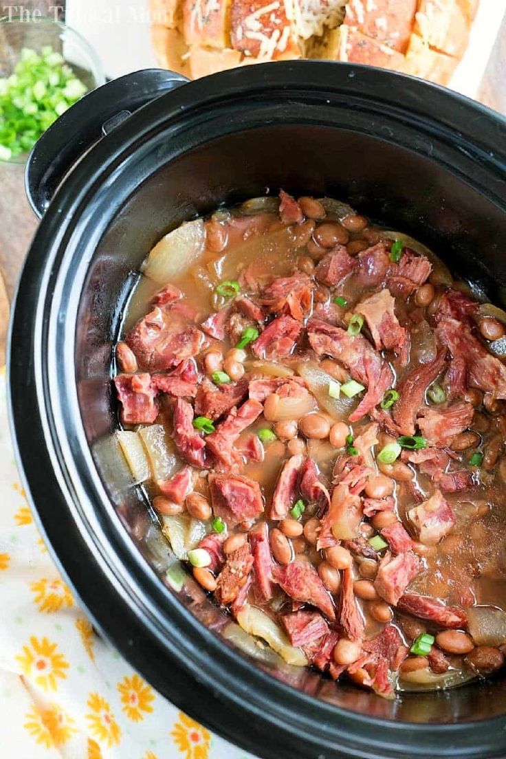 a crock pot filled with beans and meat on top of a table next to bread