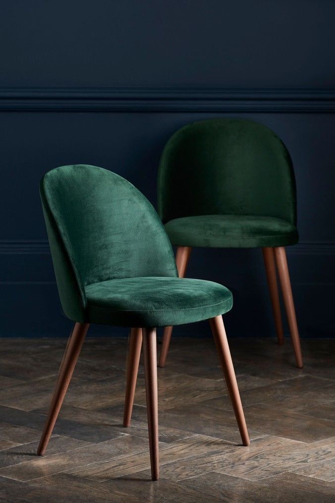 two green velvet chairs with wooden legs in front of a dark blue wall and wood floor