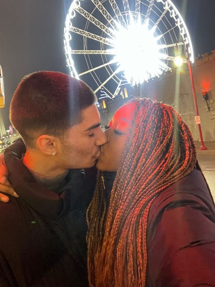 a man and woman kissing in front of a ferris wheel at night with lights behind them