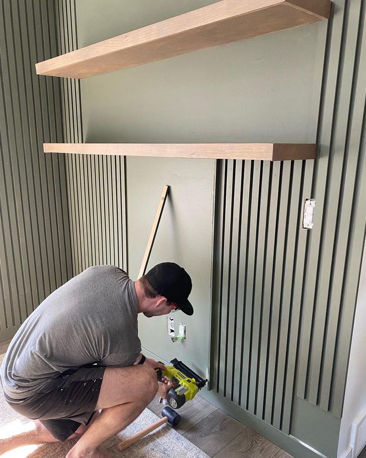 a man in grey shirt and black hat working on a wall mounted shelf with tools