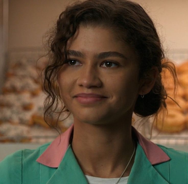 a woman with curly hair wearing a green shirt and pink collared top looking at the camera