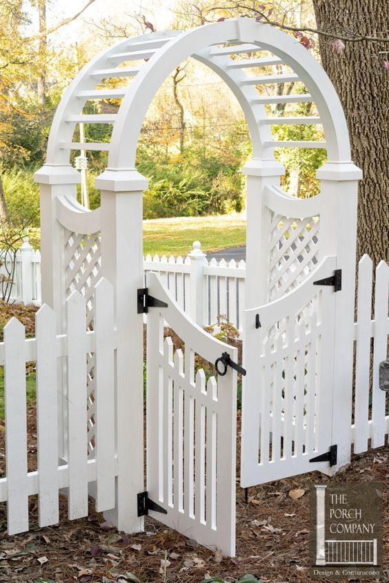 a white fence with an arched top and gate on the front side, surrounded by leaves