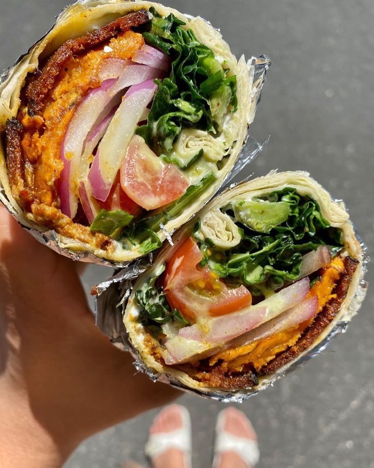 two sandwiches with meat, lettuce and tomatoes wrapped in tin foil on top of each other