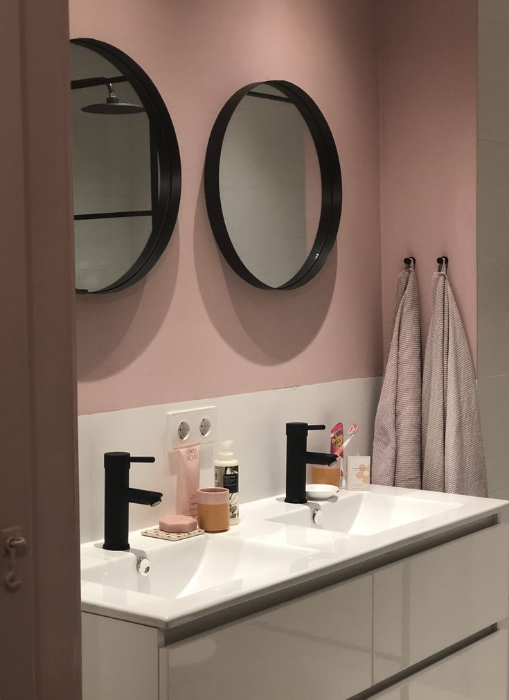 two round mirrors on the wall above sinks in a pink and white bathroom with black faucets