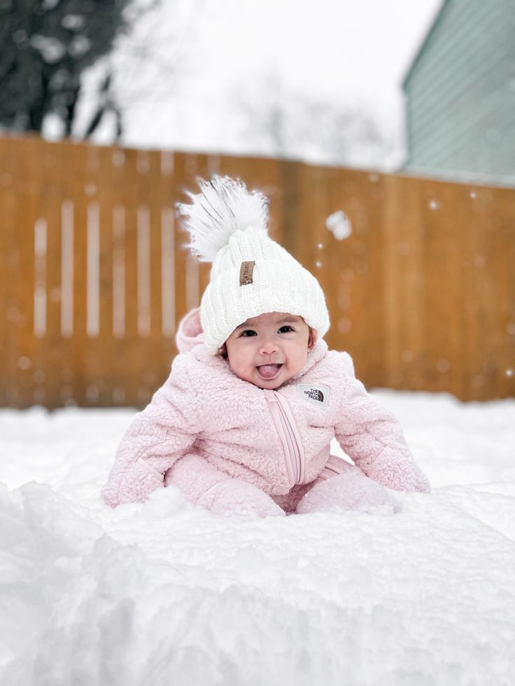 a baby is sitting in the snow wearing a pink coat and white knitted hat