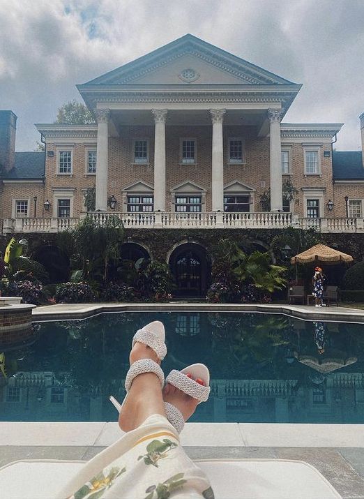 someone's feet resting on the edge of a pool in front of a large house