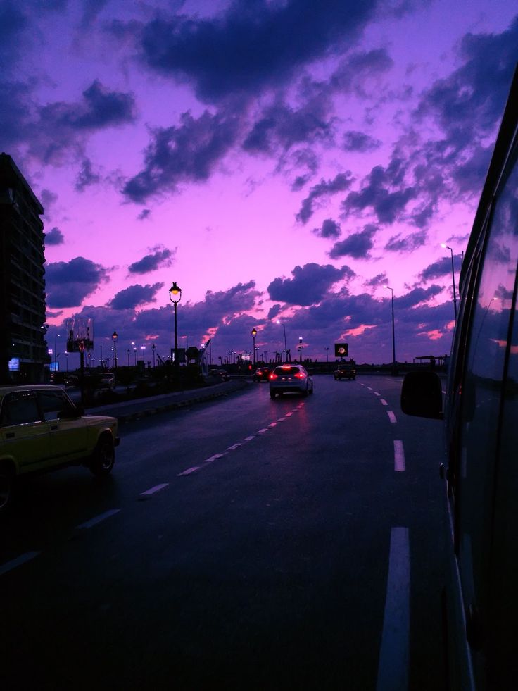 cars driving down the road at dusk with purple clouds in the sky and buildings on either side