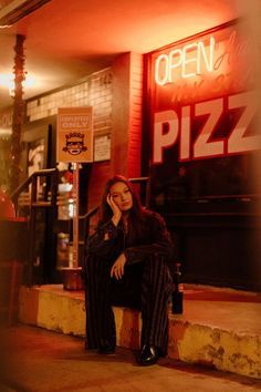 a woman is sitting on the curb in front of a pizz restaurant at night