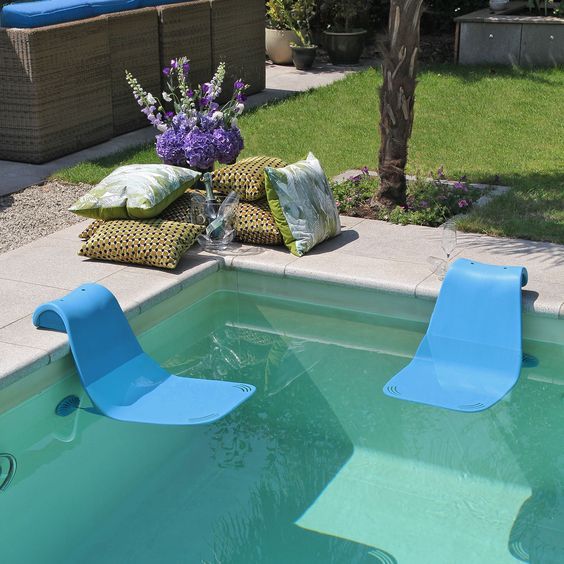 two blue lawn chairs sitting next to a swimming pool with flowers in the water and pillows on the ground