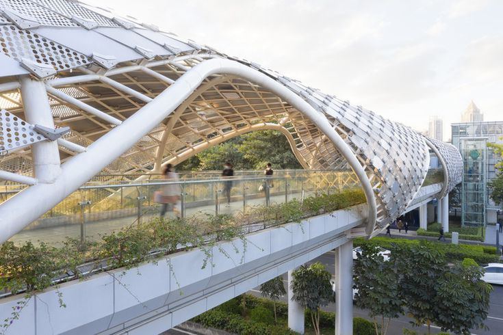 an elevated walkway with plants growing on the sides and people walking across it in front of buildings