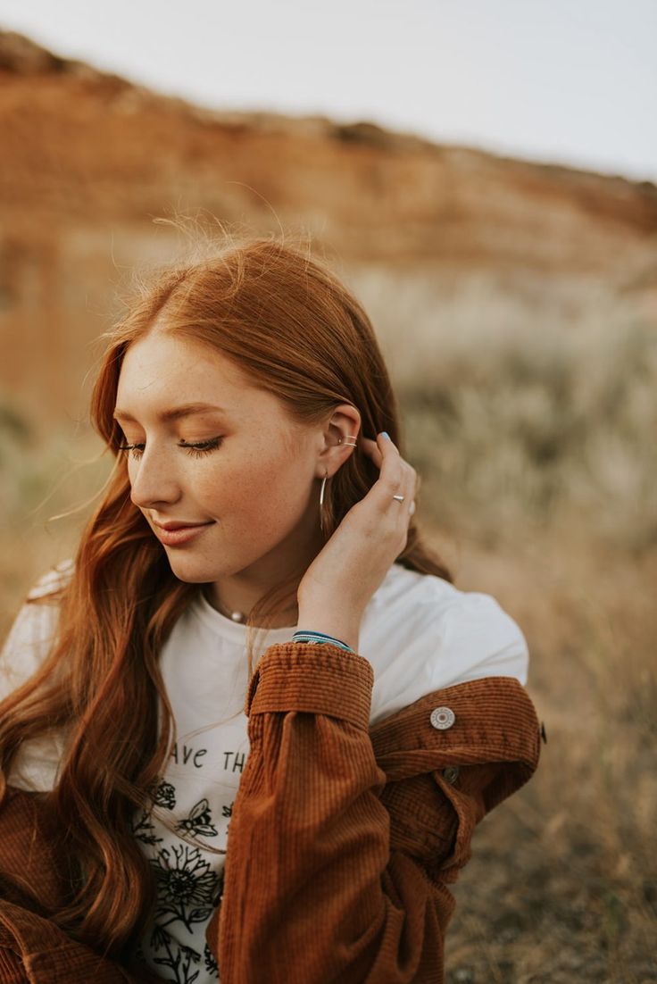 a woman with long red hair is sitting in the grass and has her hands on her ear