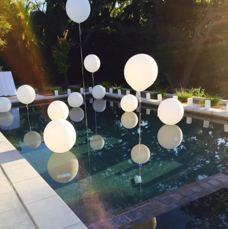 some white balloons floating in the air near a swimming pool with chairs and tables around it