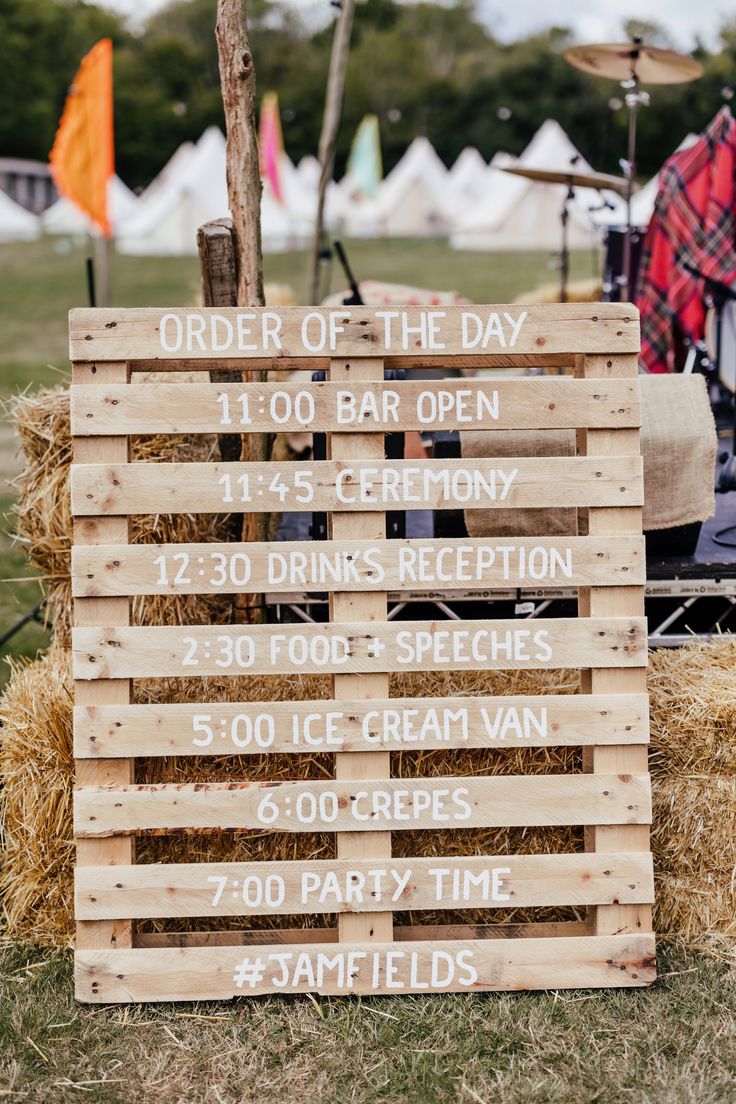 a wooden sign sitting on top of a pile of hay next to a field filled with people