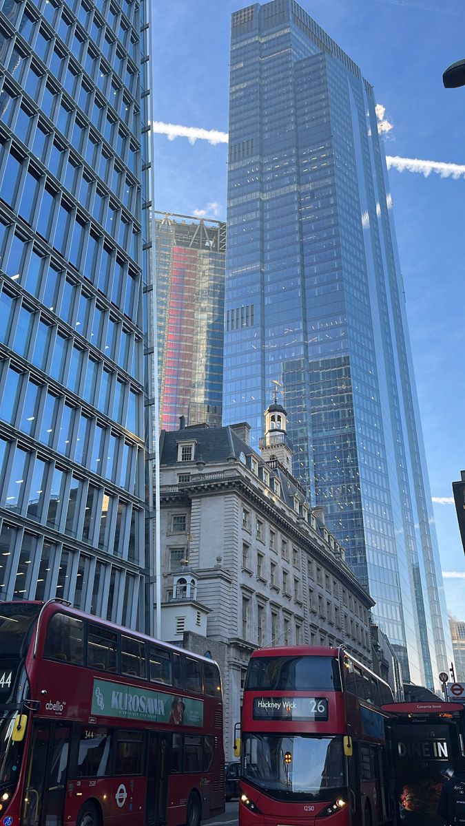 two red double decker buses driving down a street in front of tall buildings and skyscrapers