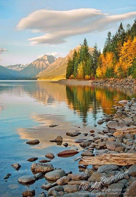 a lake with rocks and trees in the foreground, surrounded by mountains on either side