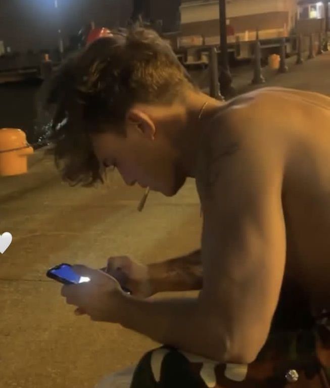 a shirtless man sitting on the ground looking at his cell phone