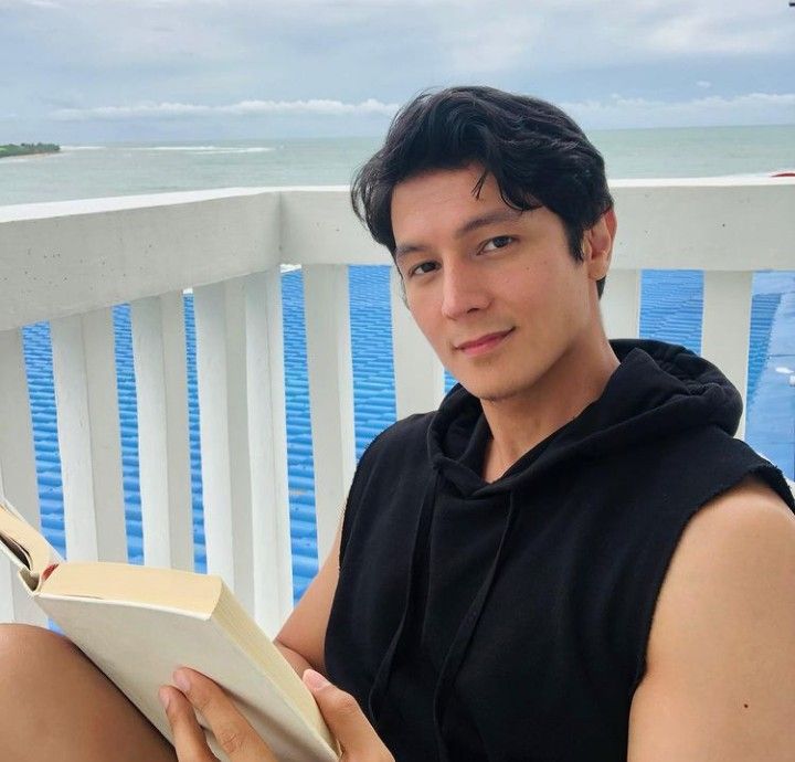 a young man sitting on a porch reading a book by the water's edge