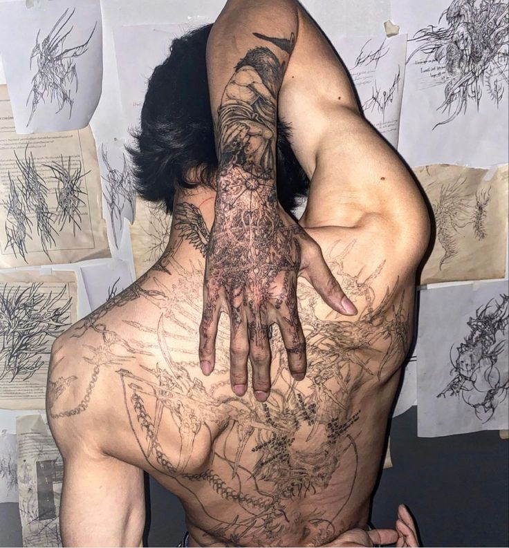 a man with tattoos on his back and hands in the air, behind him is a wall full of drawings