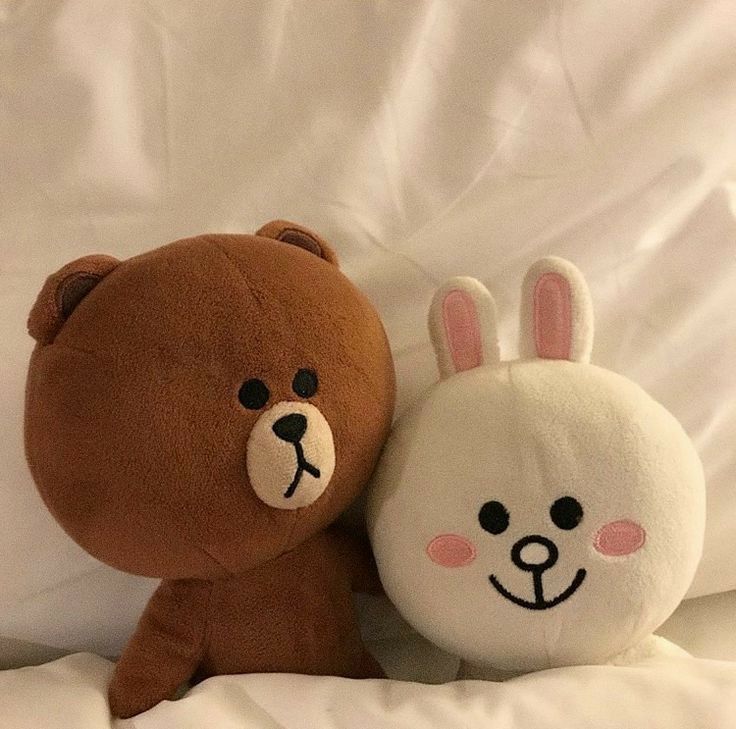 two stuffed animals sitting on top of a white bed covered in sheets and pillows, one is brown and the other is light brown