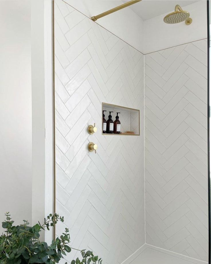 a white bathroom with herringbone tile on the shower wall and brass faucet