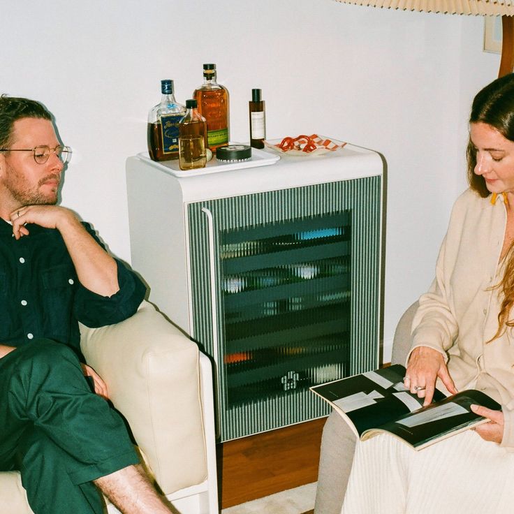 a man and woman sitting on a couch looking at an electronic device in front of a fireplace