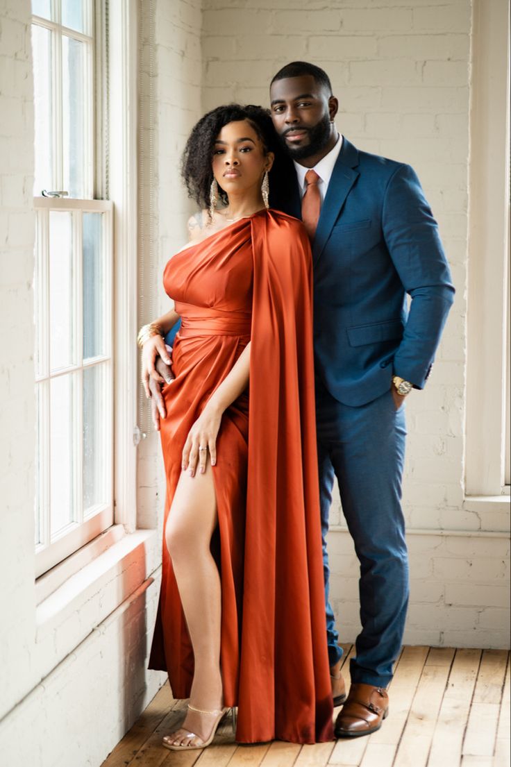 a man and woman standing next to each other in front of a window wearing orange dresses