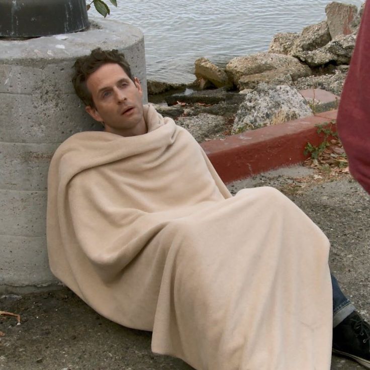 a man wrapped in a blanket sitting on the ground next to a body of water