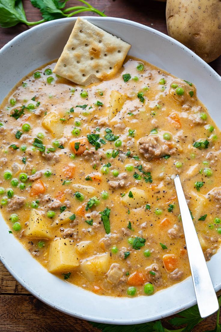 a white bowl filled with soup and crackers on top of a wooden table next to potatoes