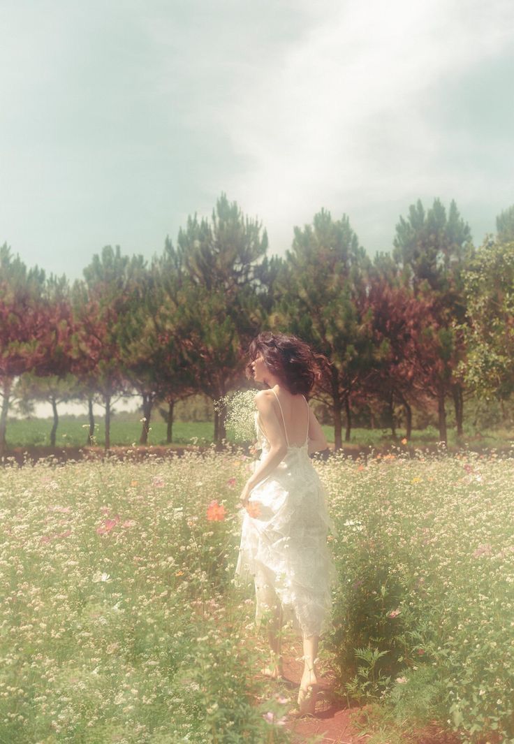 a woman in a white dress walking through a field with flowers and trees behind her