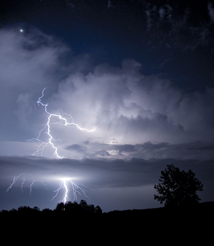 a lightning bolt is seen in the night sky over trees and hills with dark clouds