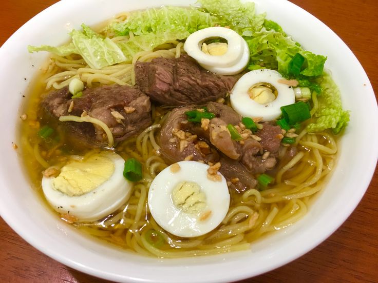 a white bowl filled with noodles, meat and boiled egg on top of lettuce