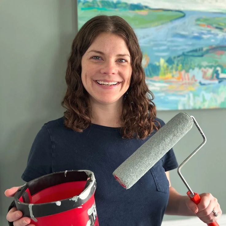 a woman holding a paint roller and a red bucket in her right hand while smiling at the camera