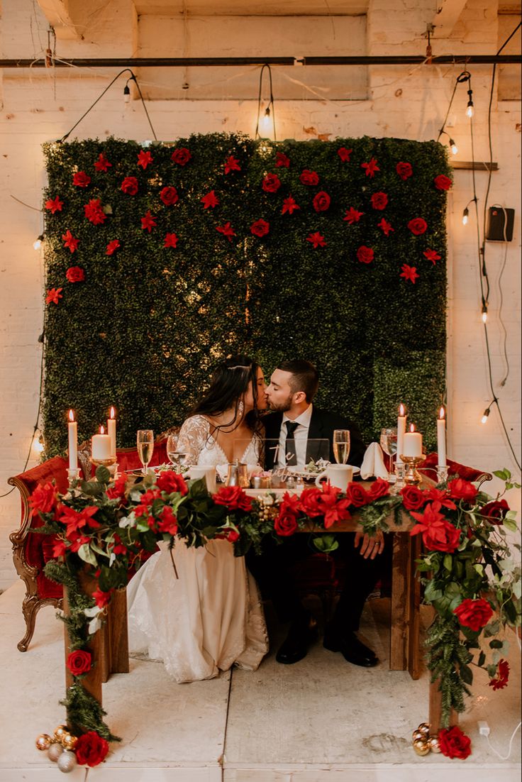 a bride and groom kissing in front of a table with red flowers on it, surrounded by candles