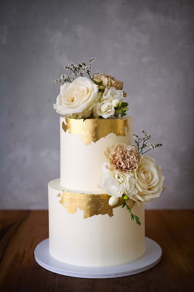 a three tiered wedding cake with white and gold flowers on the top, sitting on a wooden table